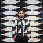 Designer Luke Haynes poses in front of his quilt - while wearing a coordinating patchwork shirt.